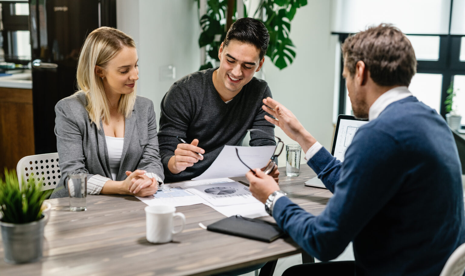 Young happy couple reading terms of mortgage documents while having meeting with real estate agent at home. Focus is on young man.